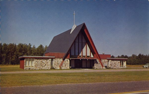Text on the reverse reads: "Shepherd of the Lakes Lutheran Church, Sayner, Wisconsin 54560. Pastor W. Wiberg." An A-frame style church with wings built of stone on each side. There is a Volkswagen automobile parked on the far right and trees fill the background.