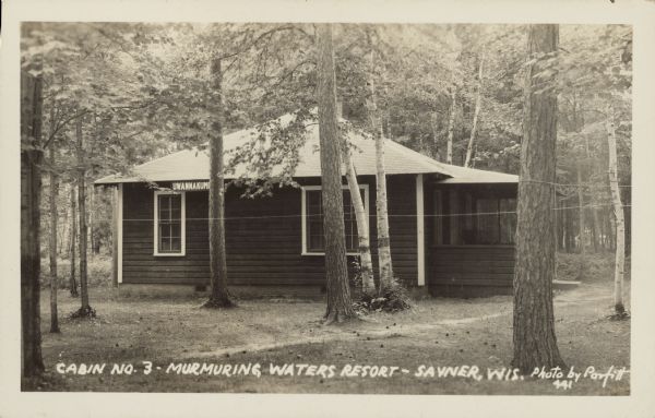 Text on front reads: "Cabin No. 3, Murmuring Waters Resort - Sayner, Wis." A clapboard cabin in the woods with a screened porch, with a path leading to the entrance. The cabin has a sign blocked by a tree that reads, in part: "Uwannakum".