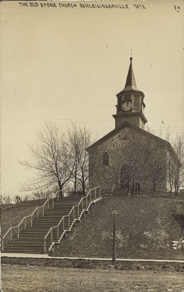 Text on front reads: "The Old Stone Church, Schleisingerville, Wis." Built around 1863, the stone church is set on a hill with a long set of steps with railings leading to the entrance. At the foot of the hill is a sidewalk and an unpaved street. The name of the town was changed to Slinger in 1921.