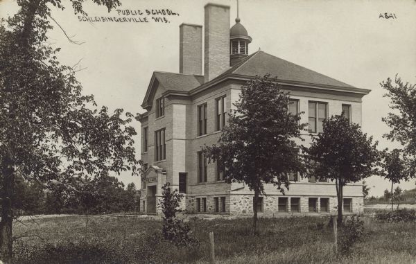 Text on front reads: "Public School, Schleisingerville, Wis." A brick and stone school building, with a belfry and two large chimneys, surrounded by trees and a lawn. The name of the town was changed to Slinger in 1921.