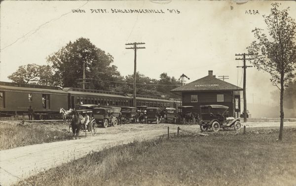 Text on front reads: "Union Depot, Schleisingerville, Wis." A railroad station with a train on the tracks. A man with a cart is standing near the baggage car on the left. There is a large group of automobiles and horse-drawn carriages near the passenger cars. Two women are sitting in a horse-drawn carriage in the center closest to the road. The name of the town was changed to Slinger in 1921.