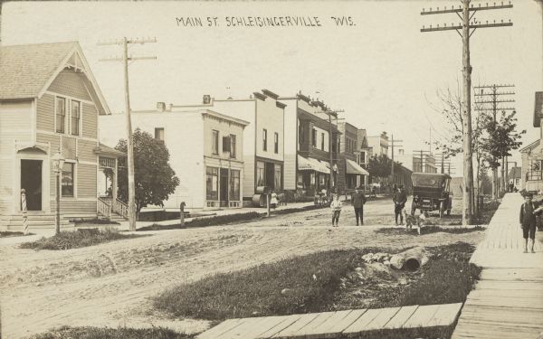 Text on front reads: "Main Street, Schleisingerville, Wis." An unpaved street lined with businesses and homes. The sidewalks are both cement and wooden boardwalks. People are standing on the sidewalks on both sides of the street in the background, and a barefoot young boy wearing a hat is standing on the sidewalk in the right foreground. Three boys and a dog are standing in the crosswalk near a parked automobile. The building across the street on the far left has a barber pole in front of the corner entrance. Further down the street on the left is what appears to be a hardware store with farm implements displayed on the sidewalk, and a furniture store. The name of the town was changed to Slinger in 1921.