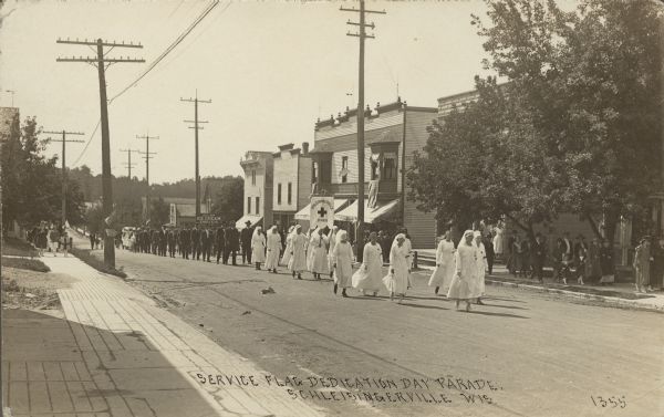 Text on front reads: "Service Flag Dedication Day Parade, Schleisingerville, Wis." Uniformed nurses are leading a parade down the street, holding a Red Cross flaf on a pole that reads: "Schleisingerville Branch". A group of men in suits are following behind. The spectators are following along on the sidewalks, with some carrying flags. Buildings, storefronts and trees line the street. In the background is a sign for "Session Ice Cream. Drugs." The name of the town was changed to Slinger in 1921.