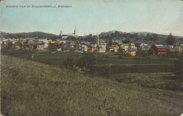 Text on front reads: "Birdseye View of Schleisingerville, Wisconsin." Elevated view from a hillside of a small town with hills on the horizon. The name of the town was changed to Slinger in 1921.