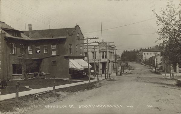 Text on front reads: "Franklin Street, Schleisingerville, Wis." View of an unpaved street with cement sidewalks in front of buildings on both sides. The large building on the left is a General Store. The awning reads: "Peter Schuck General Mdse.," Dry Goods" and "Furnishings." Farther down the block is a Millinery. The name of the town was changed to Slinger in 1921.