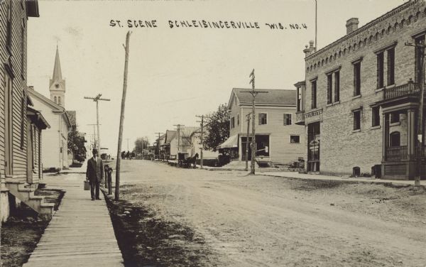 Text on front reads: "Street Scene, Schleisingerville, Wis." Buildings run along both sides of an unpaved street. One side has sidewalks, the other, a boardwalk. A man with a case is standing on the left, and several horse-drawn vehicles are in the street. In the distance is a church steeple.

The name of the town was changed to Slinger in 1921.