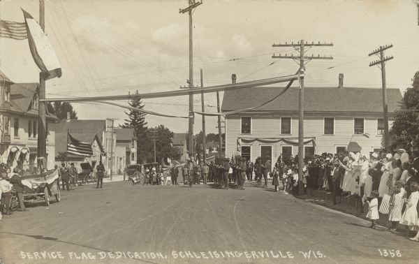 Text on front reads: "Service Flag Dedication, Schleisingerville, Wis." The people of the town and uniformed nurses are watching as the soldiers are standing at attention for the dedication of the service flag, held by a man on the left. Many people are holding American flags and the automobiles and powers poles are decorated with patriotic banners. The name of the town was changed to Slinger in 1921.