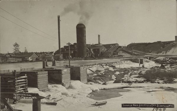 Text on front reads: "Brooks & Ross Sawmill & Dam, Schofield, Wis. 1909." The dam and millpond with the sawmill behind it. There are multiple buildings with a silo and tall chimneys.