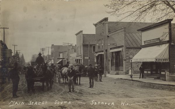 Text on front reads: "Main Street South - Seymour, Wis." Men and horse-drawn vehicles in the unpaved street with storefronts on both sides. The men are wearing work clothes. Some of the businesses include "Hoops Jewelry Co.", "First Chance Saloon" and the "Post Office". Some of the signs are in German.