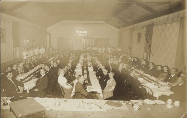 Handwritten on the back: "Business Men's Banquet, Seymour, Wisc., Thursday Eve, January 18-1912." View from stage of a large room filled with businessmen and their guests at three tables. Servers are lined up along the left wall and in the right back corner. Tableware is set along the edge of the stage in the foreground.
