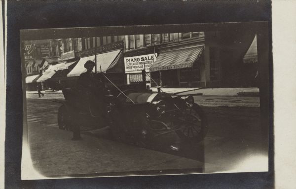 A man is sitting behind the wheel of his automobile which is parked at the curb of a street lined with businesses. A pedestrian is walking across the street in the background on the left. Signs on buildings across the street read: "Piano Sale, The Greatest Manufacturers, Sample Piano Sale", "Brunswick Lunch", "Hoffler Mfg. Co." and "Schlitz Beer, The Beer That Made Wisconsin Famous".