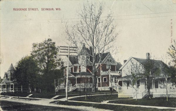 Text on front reads: "Residence Street, Seymour, Wis." A neighborhood  with the home of Dr. Hittner in the center. The street is unpaved with sidewalks and trees. Other homes are on the left and right.