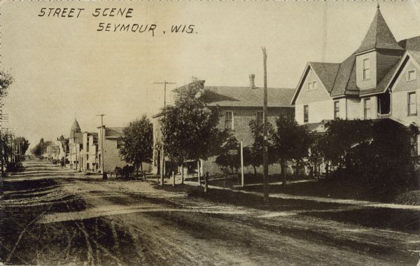 Text on front reads: Street Scene, Seymour, Wis." View across and down an unpaved street. The right side is lined with homes, businesses, sidewalks and trees. Several horse-drawn wagons are further down the street.