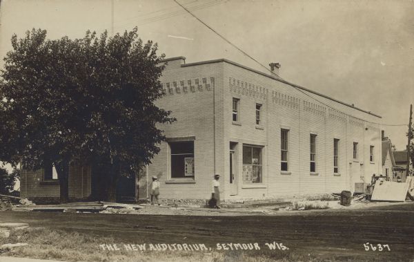 Text on front reads: "The New Auditorium, Seymour, Wis." The newly finished brick auditorium with construction debris still evident. A man and a women are walking in opposite directions on the sidewalk near two trees at the entrance. Several posters are in the windows. An unpaved road is the foreground, and two homes are on the right.<p>The Auditorium Theatre was opened around 1916. By 1941 it had been renamed See-More Theatre and continued until at least 1950.</p>