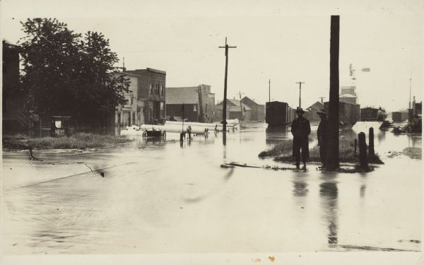 Standing water between buildings and the railroad yard. Handwriting on the back reads: "Flood at Seymoure [sic], June 3-1914." Two men are standing near the camera on the right with train cars and a grain elelvator behind them. On the left, large corrugated drainage pipes can be seen in front of the buildings.