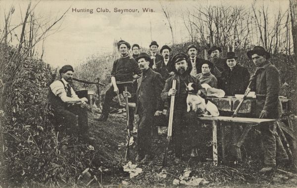 Text on front reads: "Hunting Club, Seymour, Wis." A group of men are posing with their guns and a dog around a table in a hunting camp.