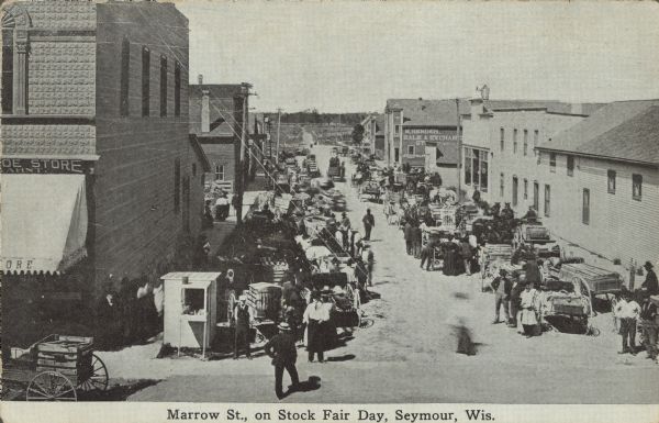 Text on front reads: "Marrow [sic] St., on Stock Fair Day, Seymour, Wis." Elevated view looking West on Morrow Street, the name is misspelled  on the front. The street is filled with buyers and sellers, wagons and animals. Businesses line the street, "Kahnt's Shoe Store" is on the left, and "M. Bender, Sale & Exchange, Stable" is on the right.