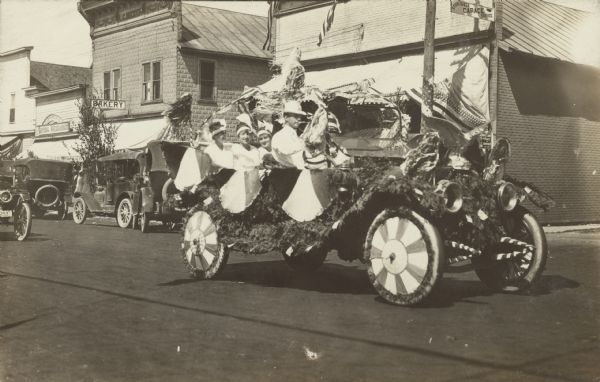 An automobile decorated for an early 4th of July parade traveling down the street. The passengers are dressed festively. Signs for "Castersen & Co. General Merchandise," "L.G. Becker Bakery" and "Studebaker Garage Service" are on the buildings in the background.