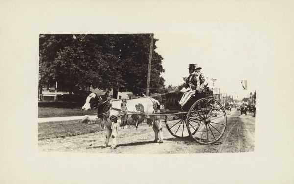 Uncle Sam and a driver in a parade buggy, pulled by a cow wearing a decorative fly net. A main street with more buggies is in the distance. American flags are hanging over the street, and a house is on the left behind trees.