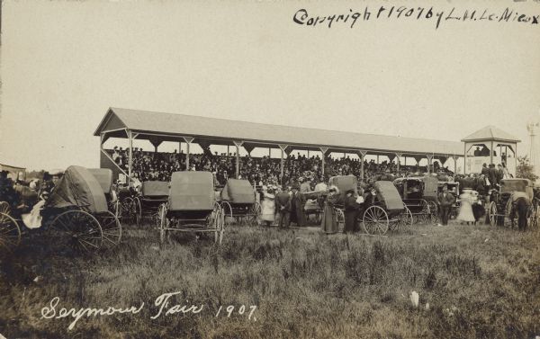 Text on front reads: "Seymour Fair 1907." The grandstand at a fair is full of spectators, with horses, buggies and automobiles parked on the infield in the foreground, with fashionably dressed people gathered near the buggies. An observation tower and windmill are on the far right. 