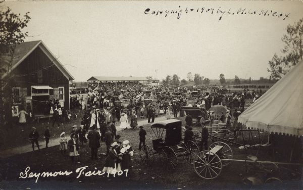 Text on front reads: "Seymour Fair 1907." Elevated view of fairgoers at the grounds with horses, buggies and automobiles. An exhibit building is on the left and a tent on the right. In the distance is a grandstand full of spectators, an observation tower and a windmill.