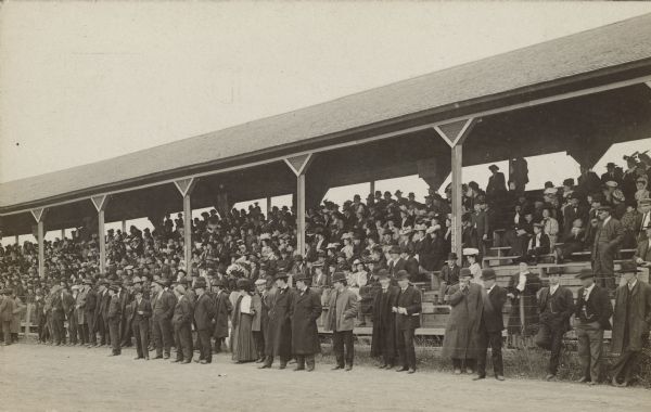 The grandstand at a fair full of spectators, and a line of people standing in the foreground at the edge of the track.