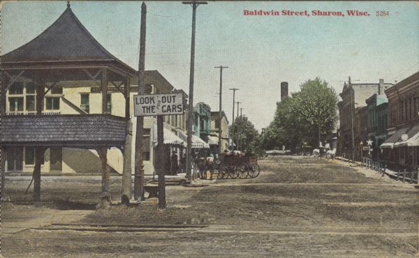 Text on front reads: "Baldwin Street, Sharon, Wisc." An unpaved street with railroad tracks crossing in the foreground. A sign near a pavilion on the left reads: "Look Out For The Cars." Several horse-drawn vehicles are hitched at the sidewalk in front of the buildings. Several pedestrians are gathered under the awning of the Hotel Sharon, on the left.