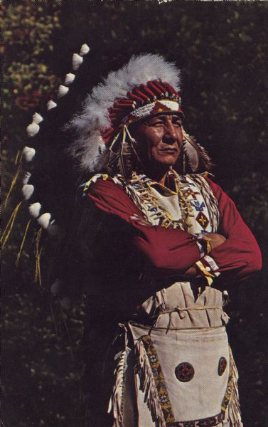 Text on reverse reads: "Menominee Chief in priceless full head dress of rare Eagle feathers. Neopit, Wis." A native American man stands with his arms crossed, garbed in indigenous dress. 