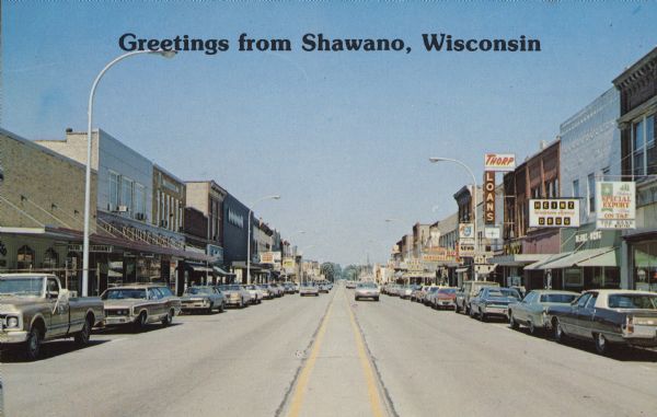 Text on front reads: "Greetings from Shawano, Wisconsin." View of South Main Street with many businesses and automobiles parked at the curb. Heinz Drug on the right was built in 1903 and is on the National and State Register of Historic Places. Other signs read: "Thorp Loans", "Schultz Bros. Co.", "Patio Restaurant" and "News Liquor".