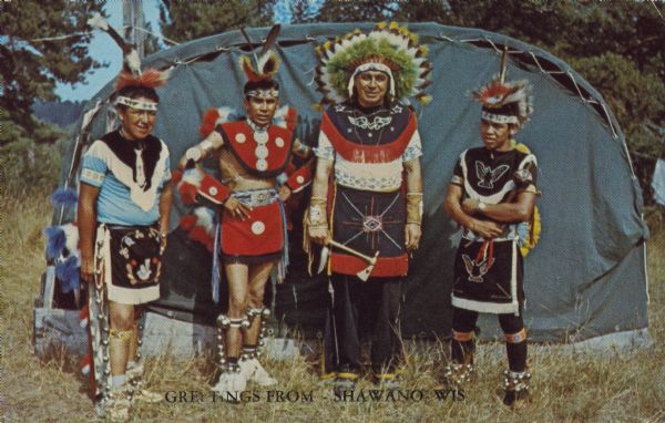 Text on front reads: "Greetings from Shawano, Wis." On reverse: "A group of Indians in their ceremonial dress." A group of Native American men are standing outdoors in front of a domed structure covered in canvas. The men are garbed in indigenous dress. 