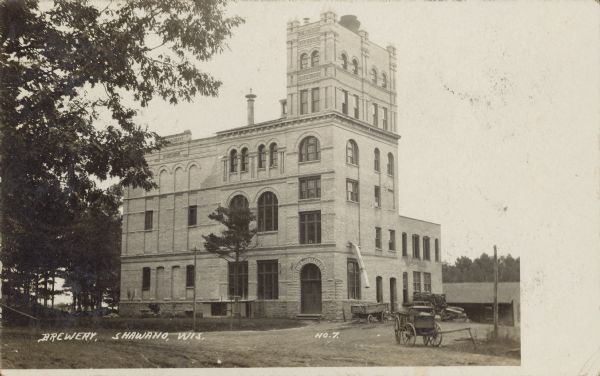 Text on front reads: "Brewery, Shawano, Wis." The second Emit T. Raddant Brewing Company building, built in 1898. The brewery is six stories tall and built of brick. It was in use until 1934 and demolished in 1967. Trees are on the left and wagons are on the right.