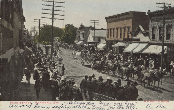 Text on front reads: "Main Street, Shawano, Wis." Slightly elevated view of a parade featuring an ornate circus wagon which is pulled by eight horses. The street is unpaved and crowds fill the boardwalks on both sides. One group is standing in a wagon for a better view. The street is lined with businesses, many with awnings.