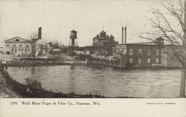 Text on the front reads: "Wolf River Paper & Fiber Co., Shawano, Wis." Industrial buildings with a millpond in the foreground. The mill was founded in 1894 and acquired by the Little Rapids Corporation in 1950. The Little Rapids Corporation still has a large operation in Shawano today.