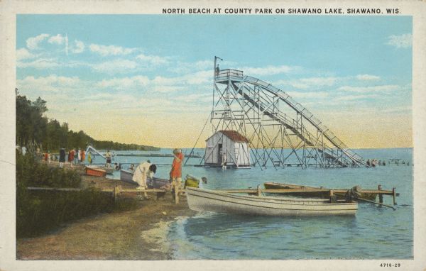 Text on front reads: "North Beach at County Park on Shawano Lake, Shawano, Wis." A giant water slide and hut can be seen in Shawano Lake at the beach. Swimmers are in the water at the end of the slide. People are standing on the sand, and piers extend into the water with row boats pulled up onto the beach. Mature trees grow on the shoreline.<p>Text on the reverse: "Six miles from Shawano, Wis., is one of the best bathing beaches in the country. That part of the beach shown in the picture is about in the middle of this 'Coney Island of Wisconsin'. The beach and park at this point are public. Maintained and operated by Shawano County. The beach is sandy, and the depth of the water increases so gradually that one-half mile from shore it is not over five feet."</p>