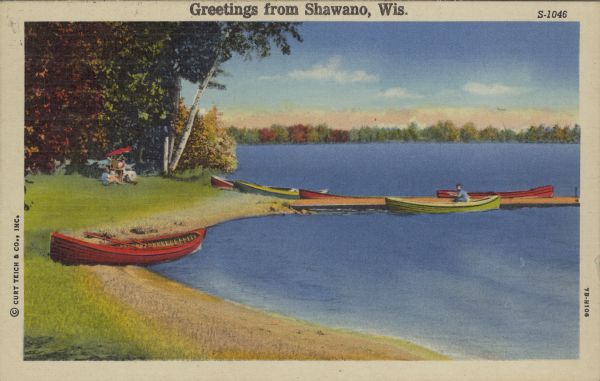 Text on front reads: "Greetings from Shawano, Wis." A scene of a beach in autumn with canoes tied up to a pier. Two people relax on the shore with a chair and umbrella, and another person is in a canoe. Colorful trees line the shore.