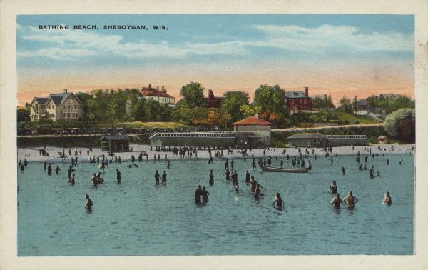 Text on front reads: "Bathing Beach, Sheboygan, Wis." View from water towards a large beach on the Lake Michigan shoreline. There is a crowd on the sand and in the water, as well as people in a boat. Several buildings are on the beach. Behind the beach is a road below a hill, with large buildings among trees in the background. Many automobiles are parked on the road.