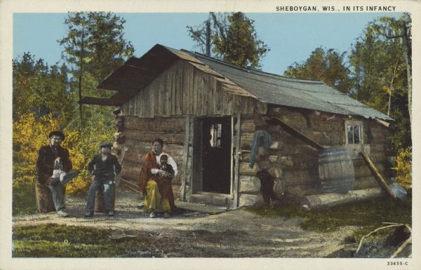 Text on front reads: "Sheboygan, Wis., In Its Infancy." A pioneer family posing in front of their log cabin, sitting on logs. The man has a dog in his lap, the women is holding an infant and a young boy is sitting in the middle. There is a rain barrel on the side of the cabin.
