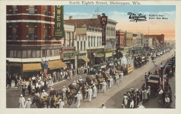 Text on front reads: "North Eighth Street, Sheboygan, Wis." There is also a Verifine Food Products logo with the text: "Equal to Any-Better than Many" in the upper right corner. A crowd on the sidewalks and in the street watch a parade of dairy themed floats and vehicles. On the left is the Wisconsin Power and Light Company and the Majestic Theater. On the marquee is "Gary Cooper, The Man from Wyoming". Automobiles are parked at the curb and American flags decorate the light posts.<p>Verifine Food Products were produced by the Sheboygan Dairy products Company, founded in 1911. It was bought by Dean Foods in 1987, then Weyauwega Cheese Company in 2016.</p>