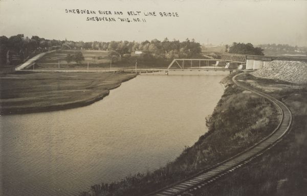 Text on front reads: "Sheboygan River and Belt Line Bridge, Sheboygan, Wis." Elevated view of a trestle bridge over the river.  Two bridges cross the river at right angles. A railroad track runs along the river on the right. Trees and buildings are in the background.