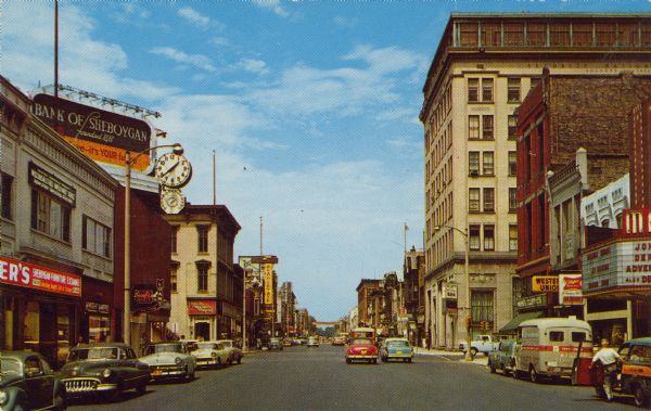 Text on reverse reads: "North 8th Street, Sheboygan, Wisconsin." View down center of a busy street lined with commercial buildings. Automobiles are parked at the curb and pedestrians are on the sidewalks. On the right is a movie theater, Western Union and a bank. On the left is a Furniture Exchange, and on the roof above is a billboard for the Bank of Sheboygan and a large clock with a thermometer. 