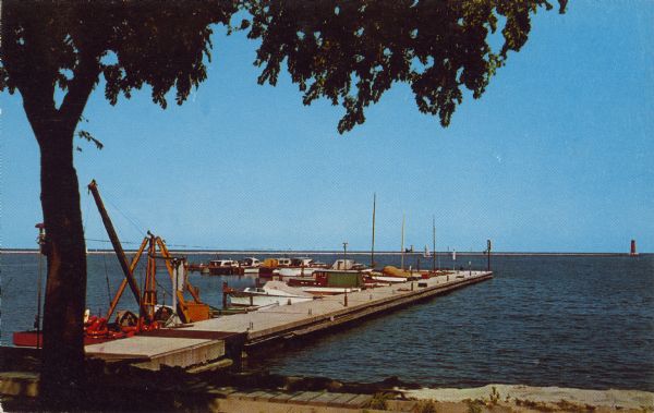 Text on reverse reads: "Yacht Basin on Lake Michigan. Sheboygan, Wisconsin." View from shore towards a long "L" shaped concrete dock with many boats moored on the shore side. A maintenance boat with a crane is docked near the shore on the left side of the dock. A large tree is in the left foreground. On the horizon is a breakwater with a lighthouse. 