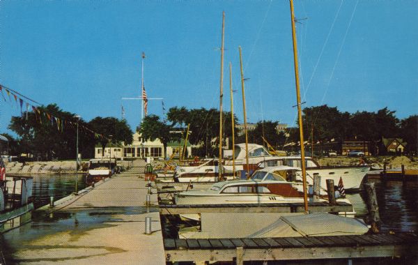 Text on front reads: "Sheboygan Yacht Club, Sheboygan, Wisconsin." View down concrete dock towards boats moored between wooden piers. In the background along the shoreline is Sheboygan Yacht Club and other buildings among trees.