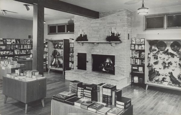 Text on reverse reads: "'Rock Ledge' Wisconsin's most picturesque gift shop located on the historical site at the Falls of the Sheboygan River, Sheboygan Falls." The gift shop has a large stone fireplace and hearth, flanked by bookcases, and display cases of land and water birds on each side. Two arrangements of pine boughs with candles decorate the mantle. Many bookcases and tables are full of books for sale. The floor is wood. The dream of Helen Stroub, it was built on four different levels that followed the contours of the rock. Rock Ledge was dedicated on October 25, 1952. Originally the site of the old Rochester Co. sawmill and the Brickner Woolen Mills warehouse, it was empty for more than 50 years. ReMax/Universal Realty Company purchased it in 2021.