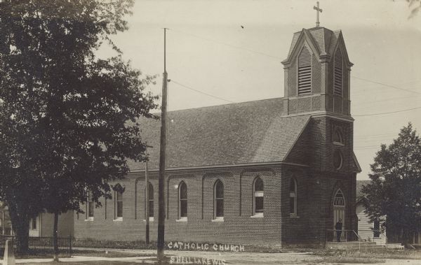 Text on front reads: "Catholic Church, Shell Lake, Wis." A brick church with a belfry and arched windows. The pastor is standing at the front entrance.