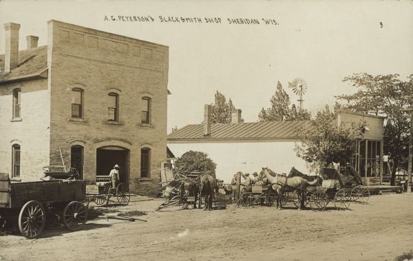 Text on front reads: "A.G. Peterson's Blacksmith Shop, Sheridan, Wis." Workers, horses, buggies and wagons are in front of a brick blacksmith's shop. The street is unpaved. Another business and a windmill are on the right.