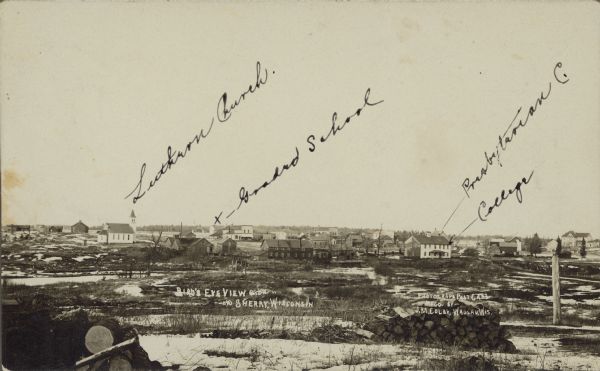 Text on front reads: "Bird's Eye View, Sherry, Wisconsin." Slightly elevated view across a field towards a small town with four locations marked in ink. "Lutheran Church", "Graded School", Presbyterian C." and "College". Two wood piles are in the foreground.