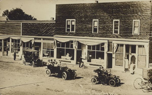 Text on front reads: "Williams Block. Shiocton, Wis." Slightly elevated view of street and commercial buildings. Two automobiles are driving past storefronts on the unpaved street. A horse-drawn buggy is on the right and a parked automobile is on the left. Several people are posing in front of the businesses. The sign on the window on the right reads: "Williams Bros."