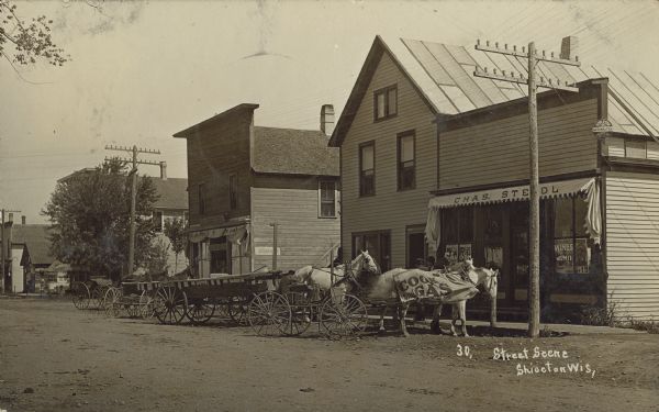 Text on front reads: "Street Scene, Shiocton, Wis." Horse-drawn wagons are hitched in front of storefronts on an unpaved street. A horse on the right wears a banner that reads: "Cook with Gas." Signs on the building on the right read: "Chas. Ste[?]dl", "Saloon", "Wines" and "Geo. Walter Beer, Appleton, Wis." There are men on the boardwalk and posters in the windows. A handwritten note on the reverse reads: "Walnut Street."