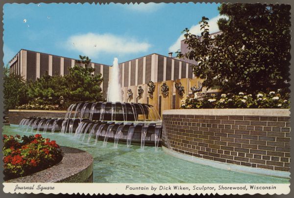 Text on front reads: "Journal Square. Fountain by Dick Wiken, Sculptor, Shorewood, Wisconsin." Text on reverse: "Journal Square. Milwaukee, Wisconsin. The home of the Milwaukee Journal, (Milwaukee's Pulitzer prize winning newspaper) and the Milwaukee Sentinal." A three part fountain cascades into a pool surrounded by several circular, flower and tree filled elements. Tall buildings are behind the fountain, and the facade of the building facing the fountain has sculptures of human figures in various poses mounted on the front. The postcard has a scalloped die cut edge.