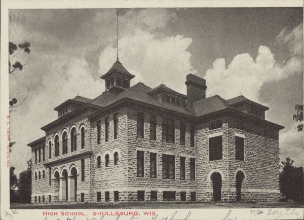 Text on front reads: "High School, Shullsburg, Wis." The Romanesque Revival school was built in 1900 of cream colored stone from the Pennich Quarry, 2.5 miles south of the city. Over the years it has been an elementary, middle, junior high and  high school.